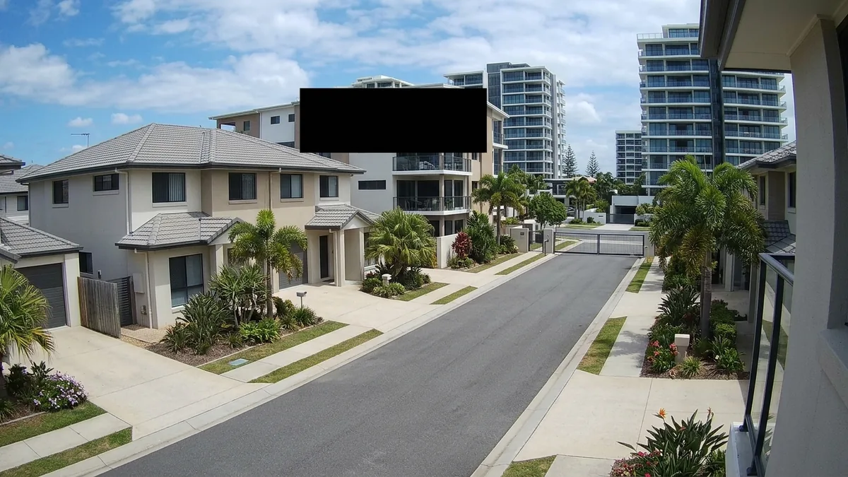 Gold Coast gated-estate CCTV view with a coastal high-rise behind and a privacy mask blacking out a neighbouring lot.